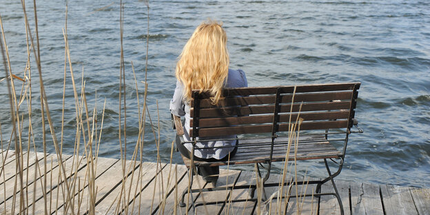 Eine Frau sitzt mit dem Rücken zur Kamera auf einer Bank auf einem Steg am Wasser