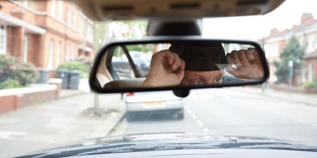 Ein Mann mit Mütze blickt vom Inneren eines Autos in einen Rückspiegel. Ein Mann mit Mütze blickt vom Inneren eines Autos in einen Rückspiegel.