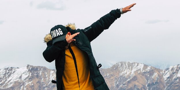 Ein Mann mit Mütze und Jacke macht vor Bergsilouette eine Tanzfigur Ein Mann mit Mütze und Jacke macht vor Bergsilouette eine Tanzfigur