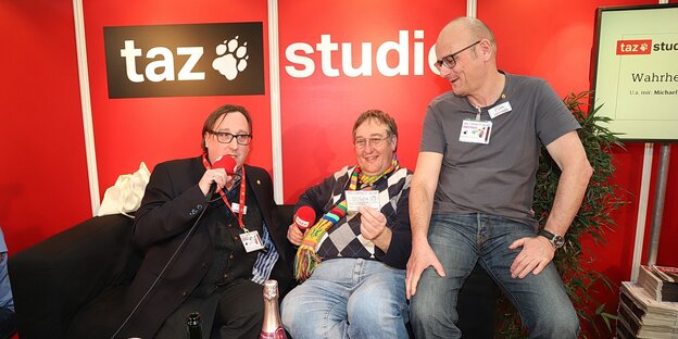 Mehrere weiße Männer sitzen vor einer roten Wand mit der Aufschrift "taz Studio" Mehrere weiße Männer sitzen vor einer roten Wand mit der Aufschrift "taz Studio"