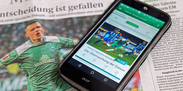Ein Handydisplay zeigt einen Beitrag der Werder-App und liegt auf einer Ausgabe des "Weser-Kuriers". Ein Handydisplay zeigt einen Beitrag der Werder-App und liegt auf einer Ausgabe des "Weser-Kuriers".