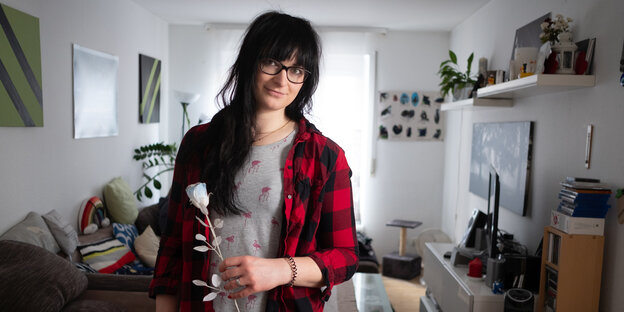Eine Person mit langen dunklen Haaren steht in einem Zimmer und hält eine weiße Rose in der Hand Eine Person mit langen dunklen Haaren steht in einem Zimmer und hält eine weiße Rose in der Hand