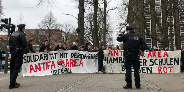 SchülerInnen demonstrieren hinter einem breiten Transparent. Im Vordergrund stehen zwei Polizisten. SchülerInnen demonstrieren hinter einem breiten Transparent. Im Vordergrund stehen zwei Polizisten.