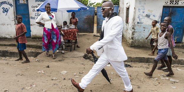 dandyhaft gekleideter Mann läuft auf einer unbefestigten Straße in Kinshasa bestaunt von Kindern und einer Frau dandyhaft gekleideter Mann läuft auf einer unbefestigten Straße in Kinshasa bestaunt von Kindern und einer Frau