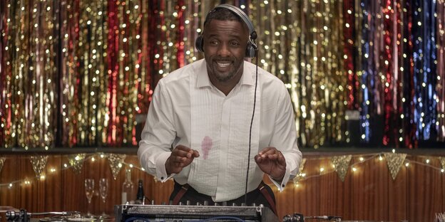 Idris Elba steht vor einem Glitzervorhang und hinter einem DJ-Pult Idris Elba steht vor einem Glitzervorhang und hinter einem DJ-Pult