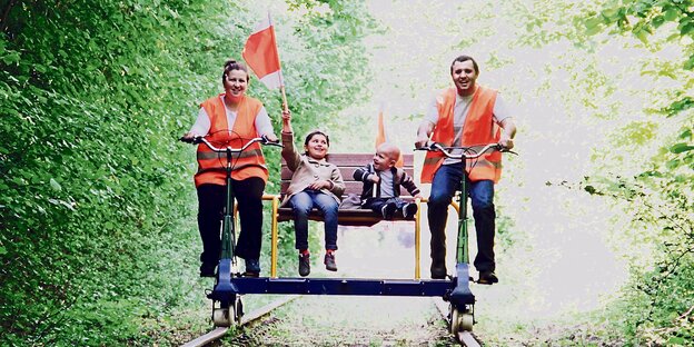 Eine Frau, ein Mann und zwei Kinder fahren auf einer Draisine durch einen Laubwald.