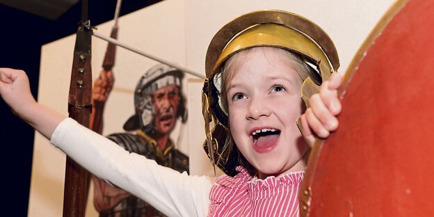 Ein Kind mit einem Legionärs-Helm steht vor der Abbildung eines Legionärs. Ein Kind mit einem Legionärs-Helm steht vor der Abbildung eines Legionärs.