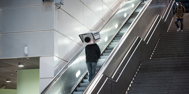 Ein teilnehmer mit einer Maske in Form einer Überwachungskamera fährt während einer Protestaktion im Bahnhof Südkreuz die Rolltreppe hoch Ein teilnehmer mit einer Maske in Form einer Überwachungskamera fährt während einer Protestaktion im Bahnhof Südkreuz die Rolltreppe hoch