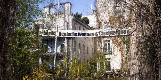 Ein Haus hinter Grünpflanzen, darauf ein Banner "Deutsche Wohnen & Co. enteignen" Ein Haus hinter Grünpflanzen, darauf ein Banner "Deutsche Wohnen & Co. enteignen"