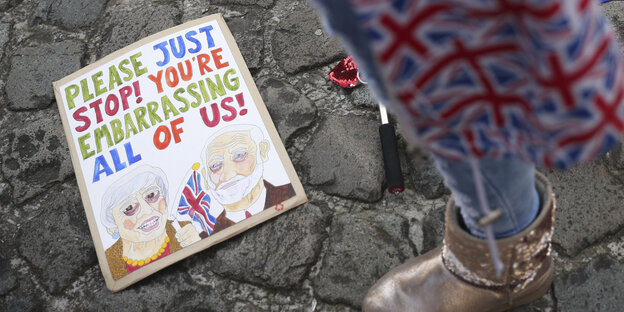 Ein Mensch steht in ein Cap mit Großbritannien-Flaggen gehüllt neben einer EU-Flagge und einem Plakat mit einer Zeichnung von Theresa May und Jeremy Corbyn und der Aufschrift: „Bitte hört auf, uns zu blamieren“ Ein Mensch steht in ein Cap mit Großbritannien-Flaggen gehüllt neben einer EU-Flagge und einem Plakat mit einer Zeichnung von Theresa May und Jeremy Corbyn und der Aufschrift: „Bitte hört auf, uns zu blamieren“