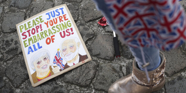 Ein Mensch steht in ein Cap mit Großbritannien-Flaggen gehüllt neben einer EU-Flagge und einem Plakat mit einer Zeichnung von Theresa May und Jeremy Corbyn und der Aufschrift: „Bitte hört auf, uns zu blamieren“