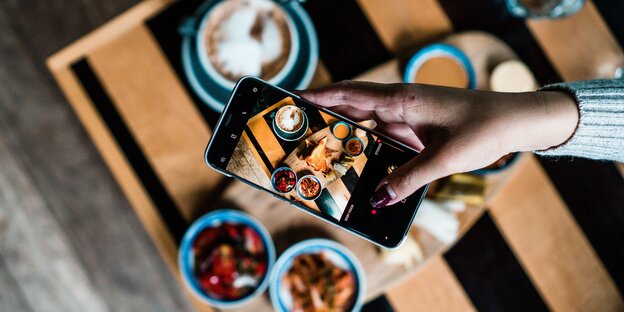 Foto eines Smartphones, das einen Tisch voller Essen fotografiert, der ebenfalls zu sehen Foto eines Smartphones, das einen Tisch voller Essen fotografiert, der ebenfalls zu sehen