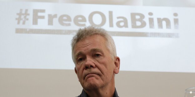 Ein älterer Mann vor einem an die Wand projezierten Bild mit der Zeile #FreeOlaBini