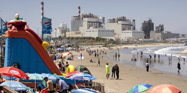 Im Vordergrund ist ein belebter Badestrand zu sehen, im Hintergrund große Fabrikgebäude