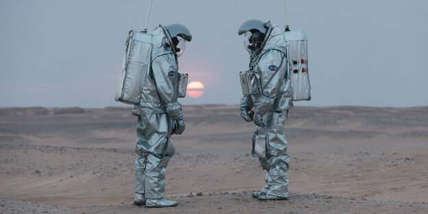 Zwei Menschen in Astronaut-Anzügen in der Wüste bei untergehender Sonne. Zwei Menschen in Astronaut-Anzügen in der Wüste bei untergehender Sonne.
