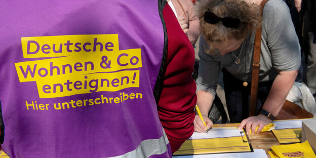 Ein mensch trägt eine Weste mit der Aufschrift „Deutsche Wohnen und Co. enteignen! Hier unterschreiben“, eine Frau stützt sich davor auf einen Tisch und unterschreibt Ein mensch trägt eine Weste mit der Aufschrift „Deutsche Wohnen und Co. enteignen! Hier unterschreiben“, eine Frau stützt sich davor auf einen Tisch und unterschreibt