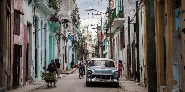 Eine Straßenszene in Havanna zeigt ein geparktes Auto, mehrere Fußgänger und bunte Häuser links und rechts Eine Straßenszene in Havanna zeigt ein geparktes Auto, mehrere Fußgänger und bunte Häuser links und rechts