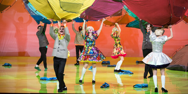 Bunt kostümierte Schauspieler halten aufgespannte Zelte über sich wie verdrehte Regenschirme Bunt kostümierte Schauspieler halten aufgespannte Zelte über sich wie verdrehte Regenschirme