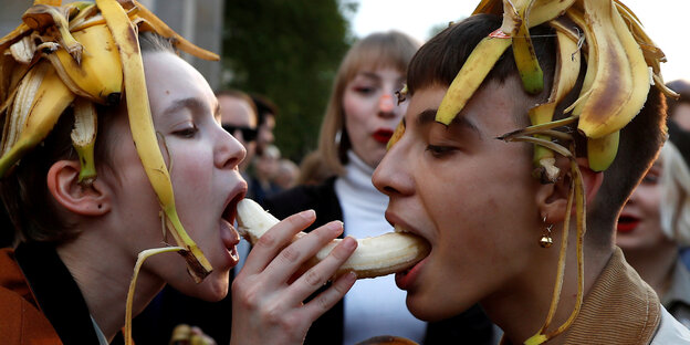 Zwei Personen haben Bananenschalen auf dem Kopf und beißen von zwei Seiten in eine geschälte Banane. Zwei Personen haben Bananenschalen auf dem Kopf und beißen von zwei Seiten in eine geschälte Banane.