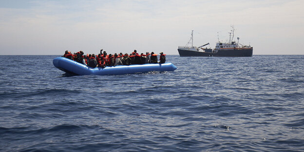 Ein Schlaucboot mit vielen Menschen, im Hintergrund nähert sich ein größeres Schiff Ein Schlaucboot mit vielen Menschen, im Hintergrund nähert sich ein größeres Schiff