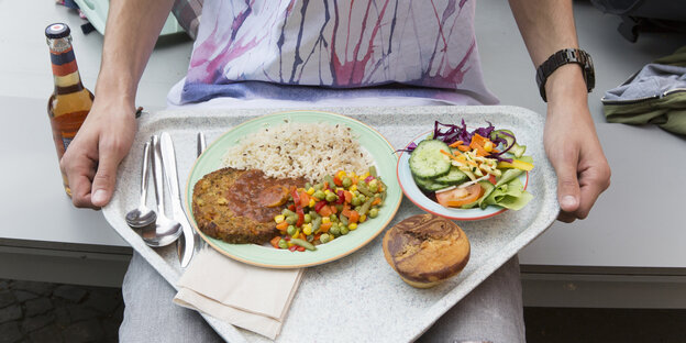 Ein Mensche trägt ein Plastiktablett mit Salat, Hauptspeise und Muffin