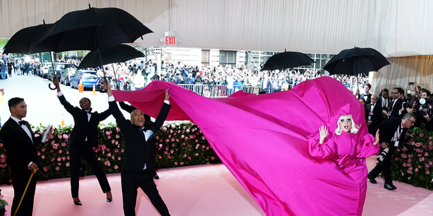Lady Gaga bei der Met Gala im meterlangen pinken Kleid umringt von schwarzgekleideten Männern mit Regenschirmen. Lady Gaga bei der Met Gala im meterlangen pinken Kleid umringt von schwarzgekleideten Männern mit Regenschirmen.