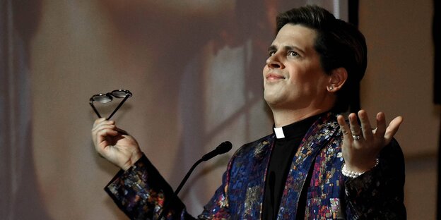 Milo Yiannopoulos hält eine Brille in der Hand und steht vor einem Rednerpult Milo Yiannopoulos hält eine Brille in der Hand und steht vor einem Rednerpult