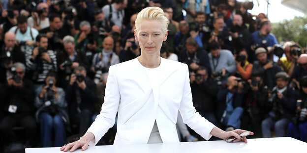 Tilda Swinton trägt einen weißen Anzug. Hinter ihr zahlreiche Fotografen Tilda Swinton trägt einen weißen Anzug. Hinter ihr zahlreiche Fotografen