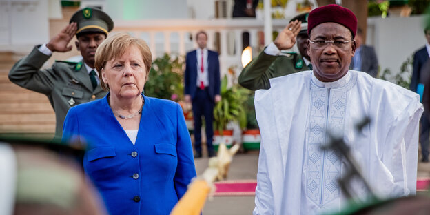 Eine Frau (Angela Merkel) steht neben einem Mann (Mahamadou Issoufou). Im Hintergrund salutiert ein Soldat Eine Frau (Angela Merkel) steht neben einem Mann (Mahamadou Issoufou). Im Hintergrund salutiert ein Soldat
