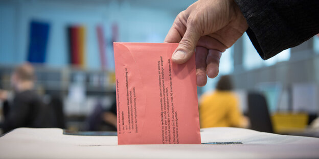 Eine Hand wirft einen roten Briefwahlumschlag in eine Urne Eine Hand wirft einen roten Briefwahlumschlag in eine Urne