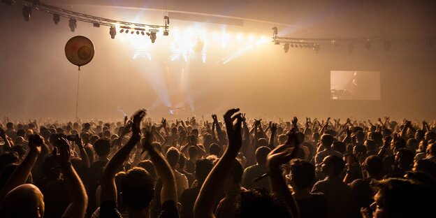 Eine große Halle mit vielen Raver*innen, in der Höhe schwebt eine Diskokugel Eine große Halle mit vielen Raver*innen, in der Höhe schwebt eine Diskokugel
