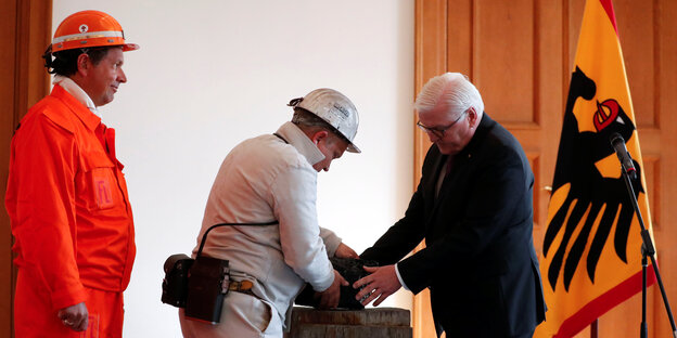 Bundespräsident Steinmeier und ein Bauarbeiter fassen zusammen einen schweren Stein an - im Hintergrund eine Deutschlandfahne Bundespräsident Steinmeier und ein Bauarbeiter fassen zusammen einen schweren Stein an - im Hintergrund eine Deutschlandfahne