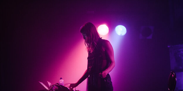 Lyra Pramuk an ihrem Synthesizer in einem Club, der mit violettem Licht erleuchtet ist Lyra Pramuk an ihrem Synthesizer in einem Club, der mit violettem Licht erleuchtet ist
