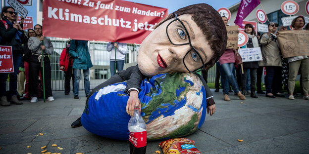 Eine Aktivistin von Campact hat eine Großmaske auf, die die CDU-Vorsitzende zeigt und liegt neben einer offenen Chipstüte und Softgetränk auf einem Sitzsack, der mit einem Erdkugel-Motiv bedruckt ist. Eine Aktivistin von Campact hat eine Großmaske auf, die die CDU-Vorsitzende zeigt und liegt neben einer offenen Chipstüte und Softgetränk auf einem Sitzsack, der mit einem Erdkugel-Motiv bedruckt ist.