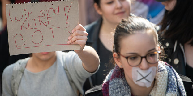 Demoteilnehmerin mit zugeklebtem Mund, hinter ihr ein Schild auf dem steht: "Wir sind keine Bots" Demoteilnehmerin mit zugeklebtem Mund, hinter ihr ein Schild auf dem steht: "Wir sind keine Bots"