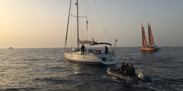 Ein Segelboot segelt in den Sonnenuntergang, dicht gefolgt von einem motorisierten Schlauchboot. Rechts im Bild sieht man ein weiteres Segelboot in der Ferne Ein Segelboot segelt in den Sonnenuntergang, dicht gefolgt von einem motorisierten Schlauchboot. Rechts im Bild sieht man ein weiteres Segelboot in der Ferne