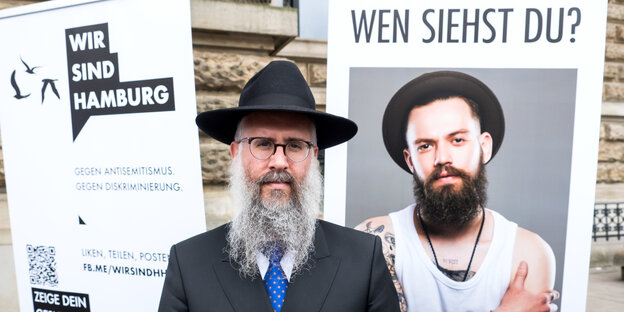 Ein Mann mit Hut und Bart neben einem Plaka, darauf: ein anderer Mann mit Hut und Bart.