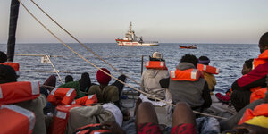 Menschen in Schwimmwesten sitzen auf einem Schiff im Mittelmeer und schauen auf ein anderes Schiff