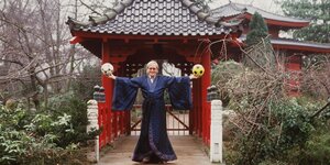 Ein Mann in einem Kimono hält je einen Ball in der Hand und steht in einem japanischen Garten