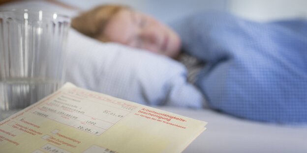 Eine Frau liegt krank im Bett, im Vordergrund ist ein Krankmeldungsschein zu sehen