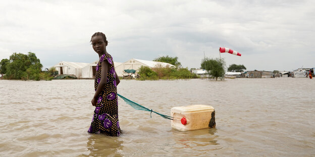 Eine junge Frau steht bis zu den Knien in dreckigem Wasser, sie zieht einen Wasserkanister an einer Leine hinter sich her