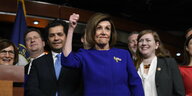 Eine Frau im blauen Blazer macht eine Siegesgeste, sie ist Nancy Pelosi, hinter ihr Männer in Anzügen