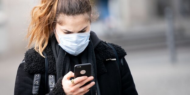 Eine Frau trägt eine Atemschutzmaske und blickt auf ihr Smartphone