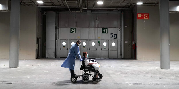Eine Person in Schutzkleidung schiebt eine Patientin in einem Rollstuhl durch eine Lagerhalle.