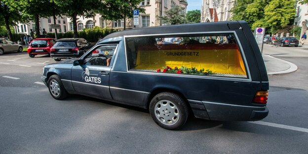 ein schwarzer Bestattungswagen, in dem liegt ein Sarg, bedeckt von einer Deutschlandfahne mit der Aufschrift „Grundgesetz“
