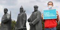 Ein junger Mann steht mit eiem Schild und Gesichtsmaske in Nowosibirsk vor drei sowjetischen Heldenstatuen