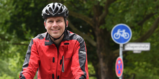 Der Antwalt Andreas Volkmann lächelnd auf seinem Fahrrad, mit Regenjacke und Fahradhelm. Im Hintergrund ein Fahrradweg-Schild. Der Antwalt Andreas Volkmann lächelnd auf seinem Fahrrad, mit Regenjacke und Fahradhelm. Im Hintergrund ein Fahrradweg-Schild.