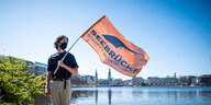 Ein Teilnehmer einer Mahnwache steht mit einer Fahne mit der Aufschrift „Seebrücke - Schafft sichere Häfen“ in Sicherheitsabstand zu den anderen Aktivisten an der Binnenalster.