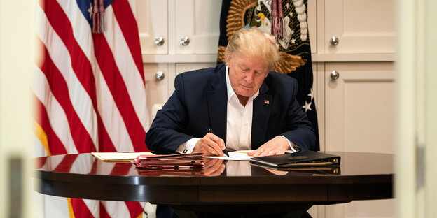Donald Trump arbeitet an einem Schreibtisch vor weißer Schrankwand und Flaggen.