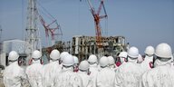 Menschen Schutzanzügen stehen vor den zerstörten Reaktorgebäuden des AKW Fukushima circa ein Jahr nach der Katastrophe